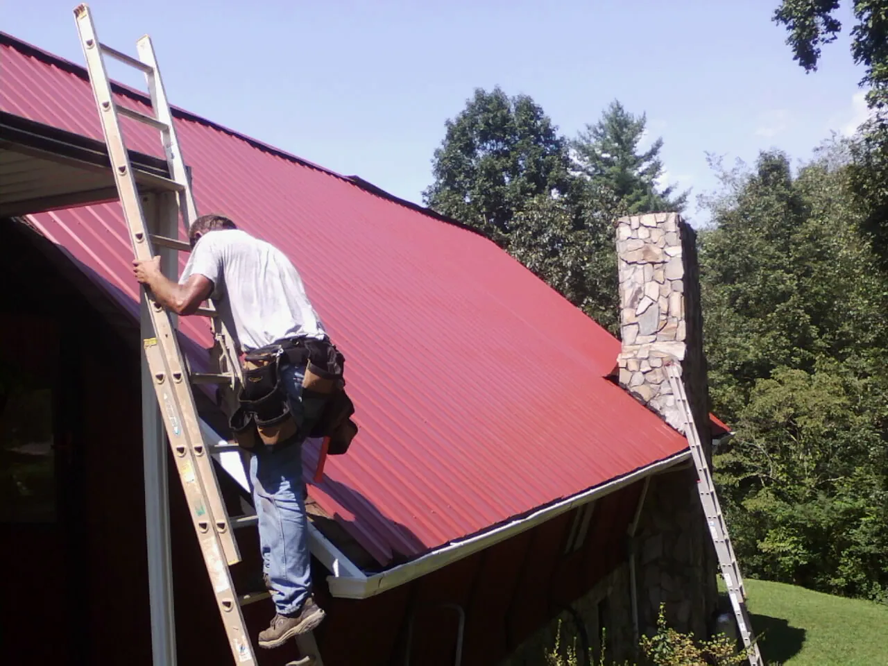 Roofer on a ladder beside a red metal roof and stone chimney — Soffit & Fascia Repair by Roofing Brothers in Tamaqua